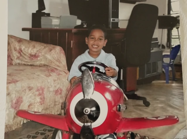 Shawn Seesahai as a child, wearing a blue shirt while sitting in an aeroplane toy and holding the steering wheel; in the room behind him is a desk, computer, and computer chair, and a sofa is to his left