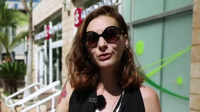 woman with light brown hair wearing black sunglasses and black top speaks to camera