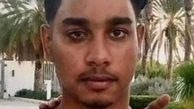 A close-up of a young man's face; he is outside and palm trees can be seen in the background