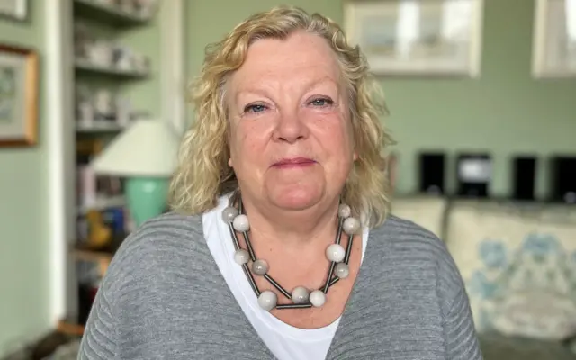 Prof Amanda Amos, wearing a grey sweater ad white t-shirt with a chunky necklace smiles at the camera in her louge