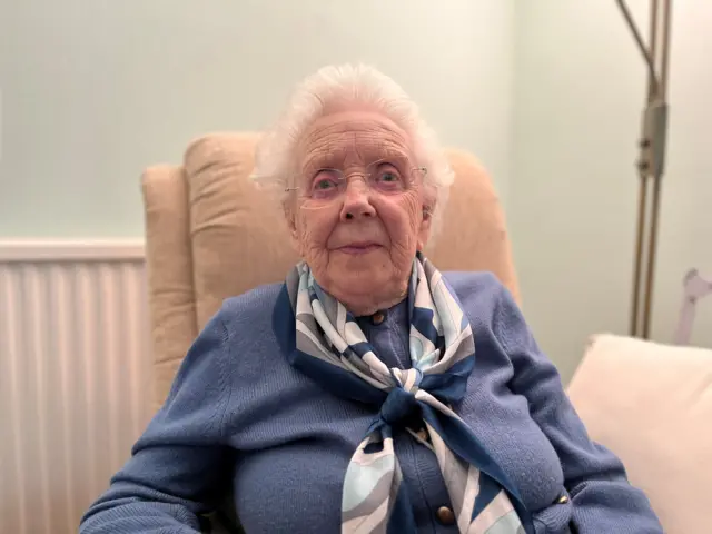 Jess Waugh is dressed smartly in a light blue cardigan and co-ordinating patterned scarf. She has white hair and glasses and looks straight at the camera while sitting in a chair beside a lamp