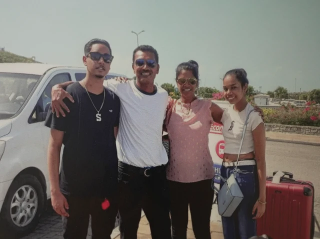 Four people stand with their arms around each other; a car is parked behind them and a red suitcase is to their right. They are standing outside and everyone is smiling.