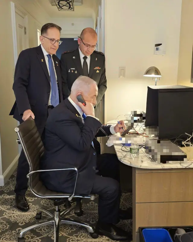 Benjamin Netanyahu sat at a desk using a landline phone, several devices on the desk have been pixellated. An official in a suit and a military officer stand behind him