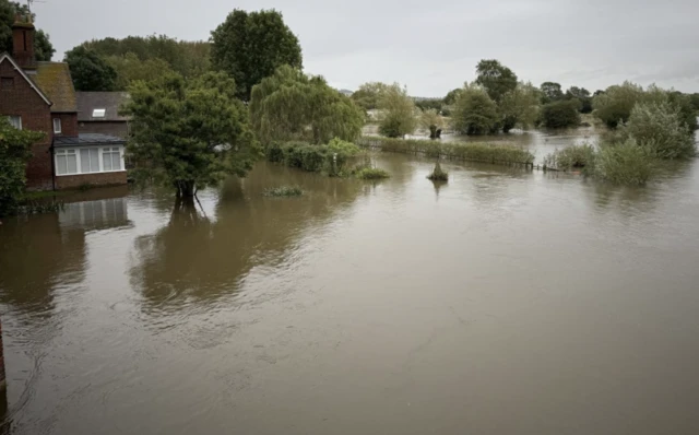 A general view of flooding in Abingdon, Oxfordshire, The image shows brown rain water up to hedges and the trunks of trees. A house in the distance can also be seen, although the water is not as high as the windows