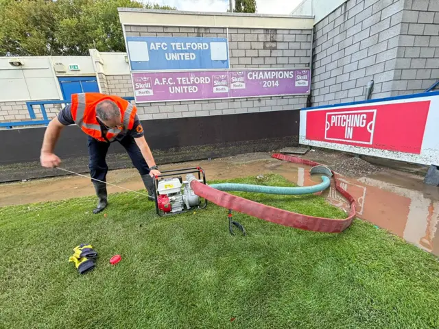 A man in high is using a pump to remove flood water from the football ground