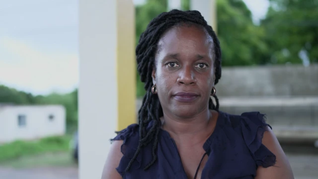 A woman with dark hair and a dark blue top looks at the camera