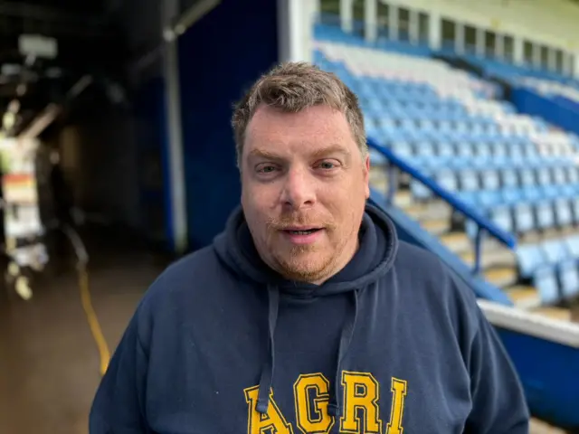 Kevin Short in a blue hoodie standing in front of seating at AFC Telford