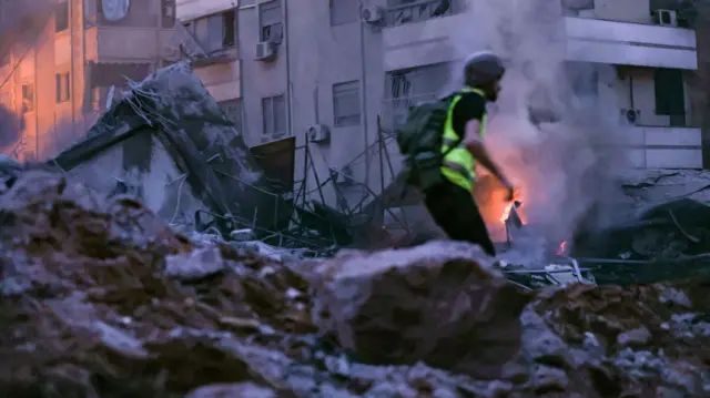 A rescue worker runs amid the rubble of a building destroyed in an Israeli air strike, there is smoke and flames in the background