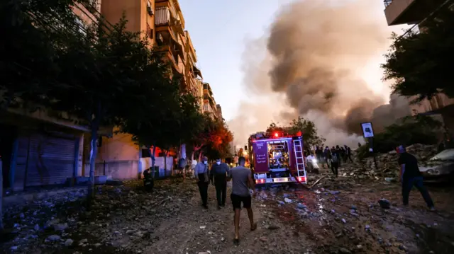 People and a fire truck rush to the scene of an Israeli air strike, a large smoke billows in the background