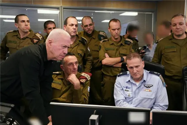 Israeli defence minister Yoav Gallant at Israel's air force headquarters during a strike on southern Beirut. Eight men in uniform around him, all looking at some screens. Several faces in background blurred out.