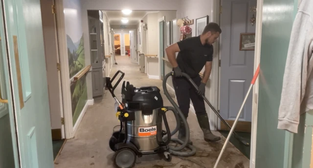 A man hoovering a corridor of the care home. He is wearing a black top and trousers