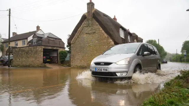 A small silver car drives through floodwaters past thatched-roofed homes