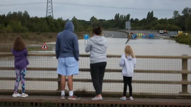 A family of four stand on an overpass looking at workers draining water from the dual carriageway, which is severely flooded