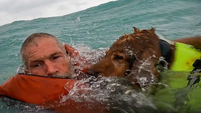 A man and his dog, both in life jackets, being rescued by the US Coast Guard