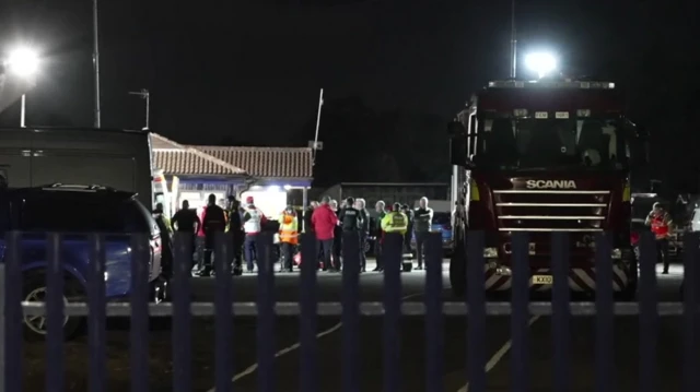 Taken from afar, dozens of people including firefighters stand outside a building during at nighttime