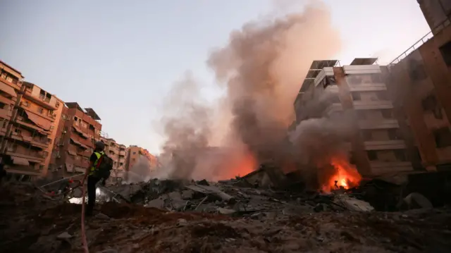 A rescue worker runs amid the rubble of a building destroyed in an Israeli air strike, there is smoke and flames in the background