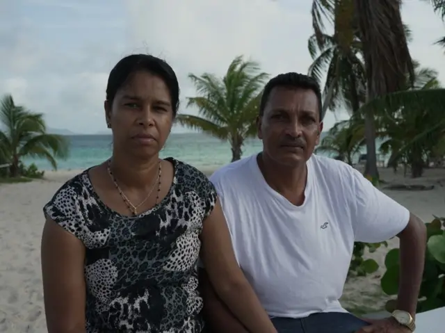 A woman and a man stare at the camera with serious faces; they are on a beach with palm trees and the sea is visible