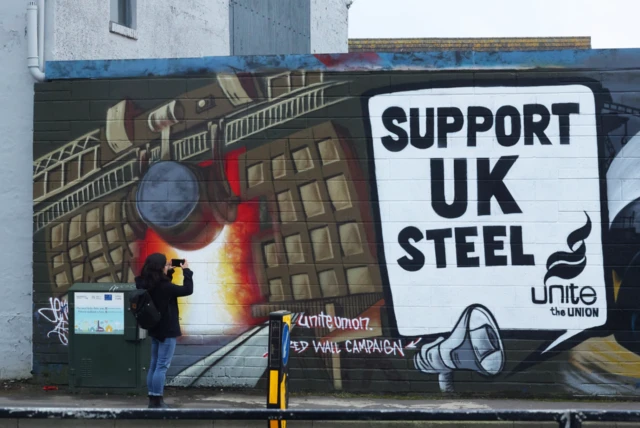 A person photographs a mural which says Support UK Steel, Unite the union, near the Tata Steel Port Talbot steel production plant