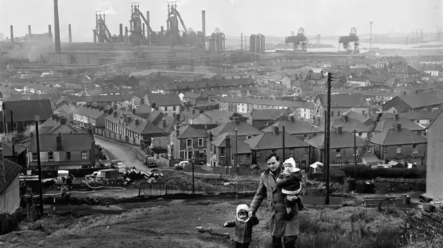 A man and two children going up a hill in Port Talbot, with houses and the steelworks behind them, in this black and white photo from the 1960s