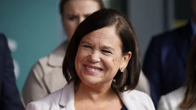 Mary Lou smiling to the side. She is wearing silver drop down earrings and a cream blazer. She has short to shoulder hair in a middle parting.