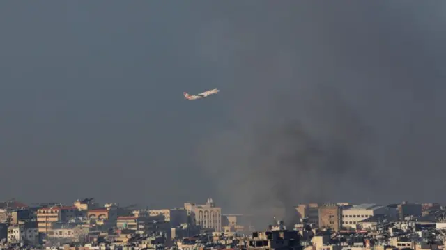 An aeroplane flies over the skyline of Beirut as smoke billows upwards