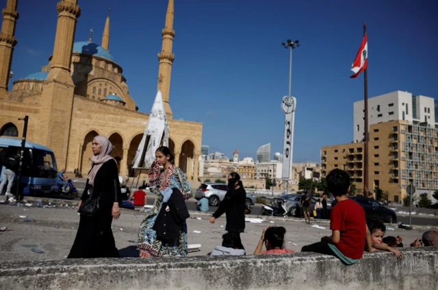 Families gatehr at Martyr's Square in Beirut with debris and rubbish lying around the place. A child wearing a red shirt has his back to the camera as he sits on a wall looking up towards a Lebanese flag which flutters in the wind