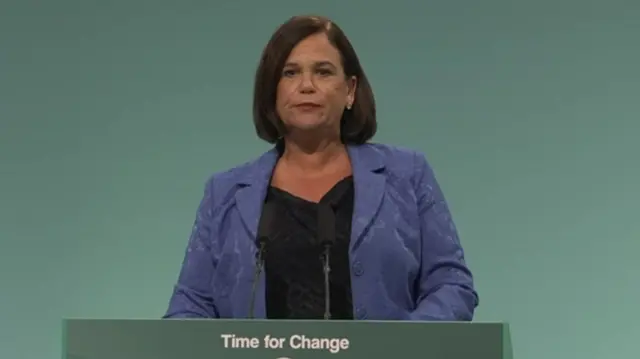 Mary Lou is wearing a purple blazer and black top. She is addressing conference at a green podium that says ''Time for change' on it