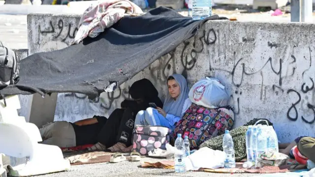 Several people sitting on the ground with a makeshift roof made from a blanket, surrounded by bags with their belongings and bottles of water