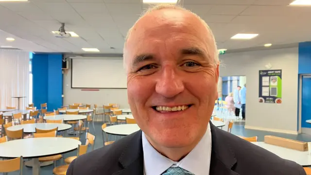 A man smiling, he has white hair and is wearing a suit