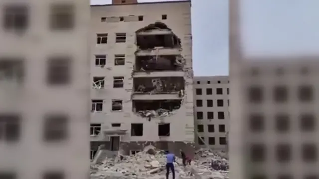 Rubble at the bottom of a multi-storey building with no glass windows and a big hole exposing floors on the side of the building