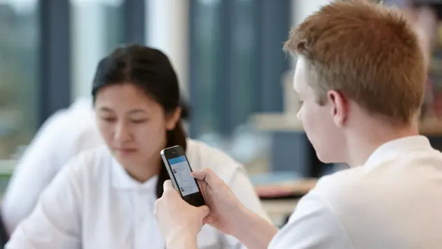 Teenage boy using mobile phone in classroom. Teenage girl sitting in background