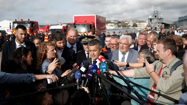 Gérald Darmanin, speaking outside with emergency vehicles in the background, addresses a crowd with several microphones in front of him