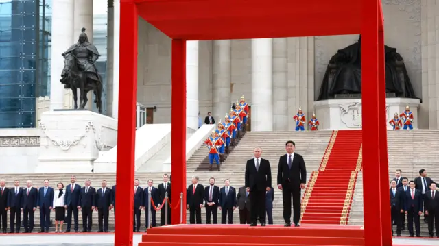 President Ukhnaagiin Khurelsukh and President Vladimir Putin stand on a red, raised platform in a big red cube structure. People in suits stand in a row behind them and there is a red carpet going down steps leading to the raised platform.