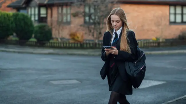 A young girl in a school uniform walking along street looking at a mobile phone