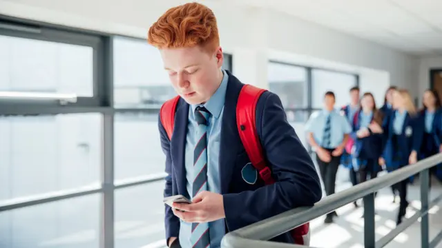 A pupil with red hair wearing a navy blazer and blue shirt, they are on their phone