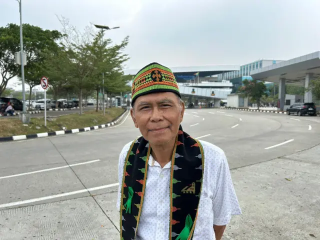 A man in a hat and scarf standing outside an airport in a white button-up shirt