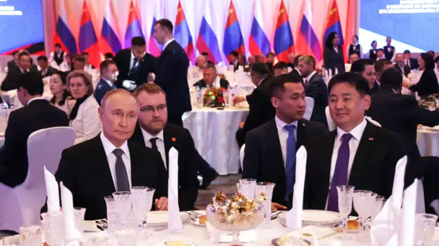 President Vladimir Putin and President Ukhnaagiin Khurelsukh sit at a table with white clothe and crockery on it. There are many people in suit on similar round white tables behind them