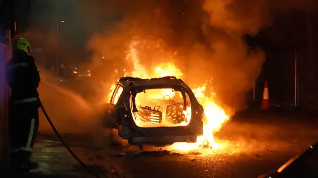 A car on fire with a fireman using a hose to try and put it out