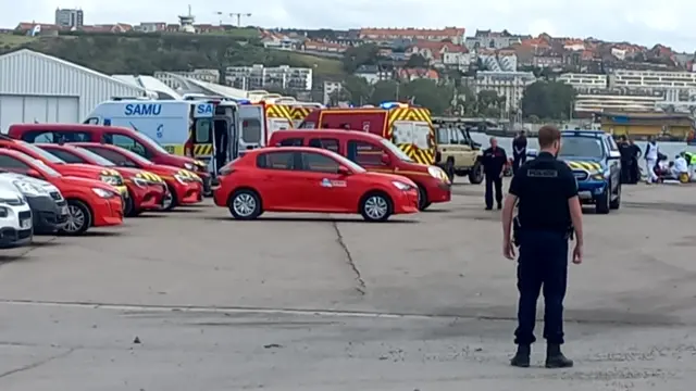 A police officer stands in the foreground with emergency vehicles in the backgroun