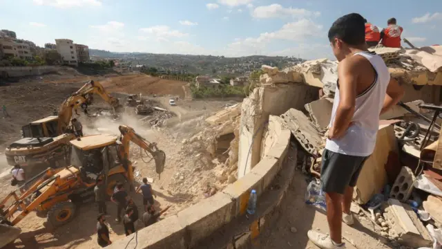 Man looks down on pile of rubble in Ain al-Delb as diggers work to clean up rubble