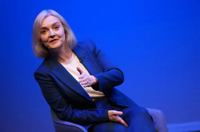 Liz Truss sitting down on chair on stage wearing royal blue suit and white shirt