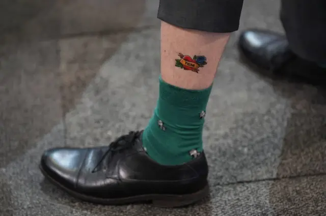 Close up of man's left foot sporting a temporary calf tattoo in the shape of a heart and the name Tom