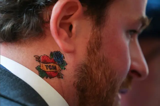 heart-shaped temporary tattoo with blue roses and Tom script on nech of bearded man in suit