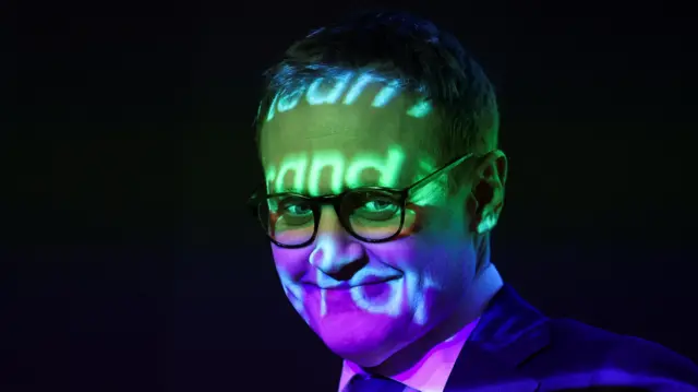 Conservative Party MP Tom Tugendhat, his face is illuminated by a green light in the top half and a purple one in the bottom. Some text is also visible, but not legible.