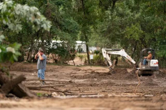 A man talks on his cell phone while a tractor does work nearby