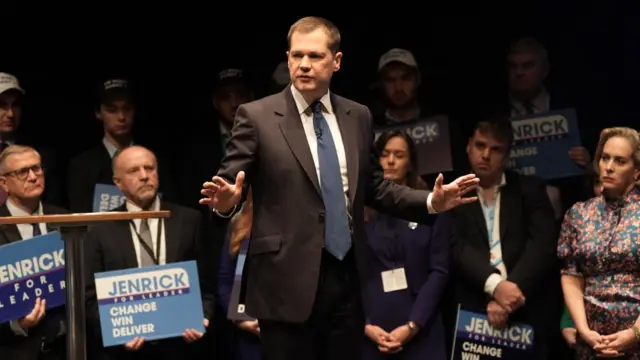 Robert Jenrick on stage at a rally for supporters. People behind him are holding signs which say "Jenrick for Leader: Change, Win, Deliver"