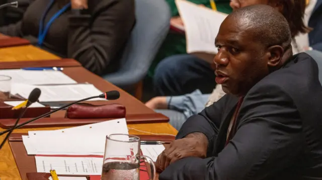 David Lammy sits at a desk with a microphone and papers on it