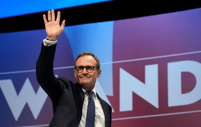 tugendhat waves at conference attendees as he walks on stage wearing dark suit and dark blue tie