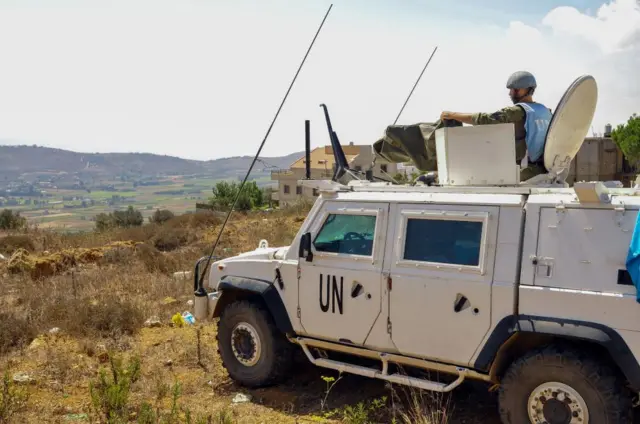 A white truck overlooking a hilly area in southern Lebanon. Truck has a UN sign on the door, and a peacekeeper with a blue vest and helmet, standing up in the hatch