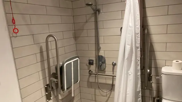 A shower in a bathroom. The tiles are white, as is the shower curtain, which is drawn back.